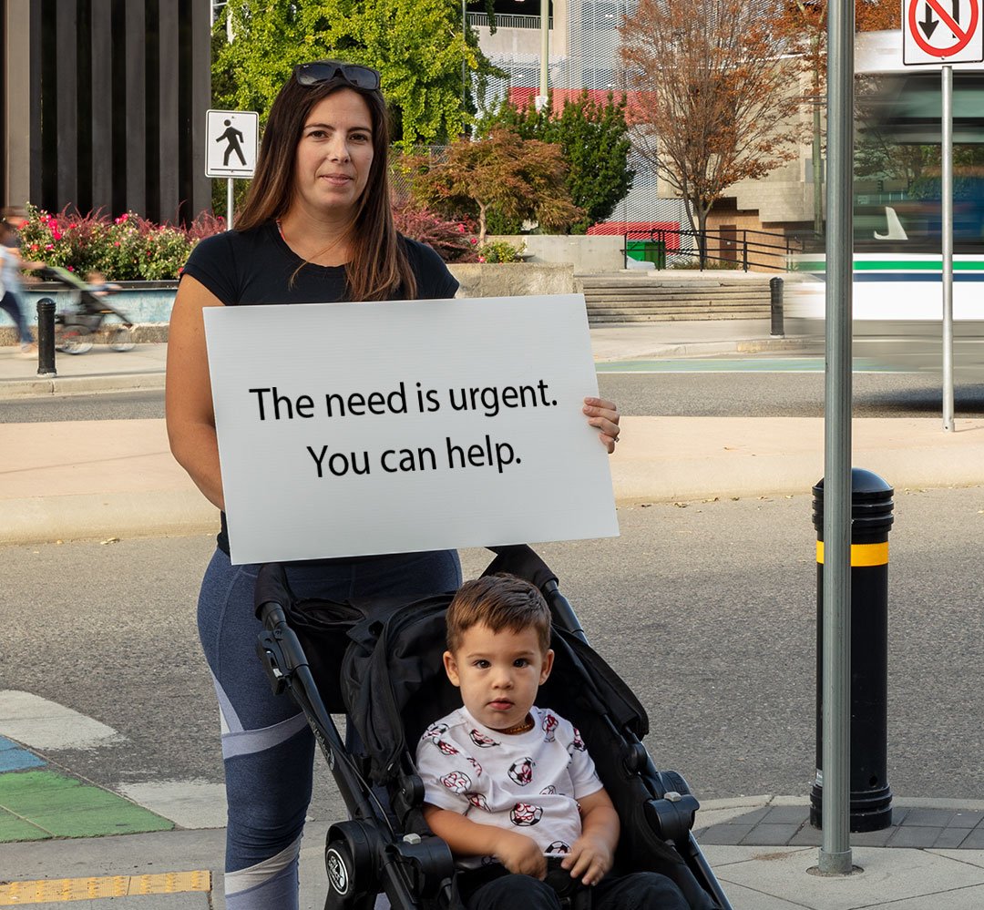 Y Give (teaser) Mother holding sign that says: The need is urgent. You can help.