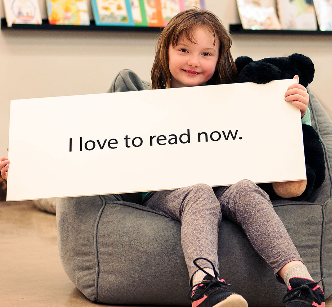 AR 2021 BTB Teaser 7 year old girl smiling and sitting in a grey bean bag chair with a teddy bear holding a large white sign the reads 'I love to read now'. There is a library/ classroom background with shelves of books.