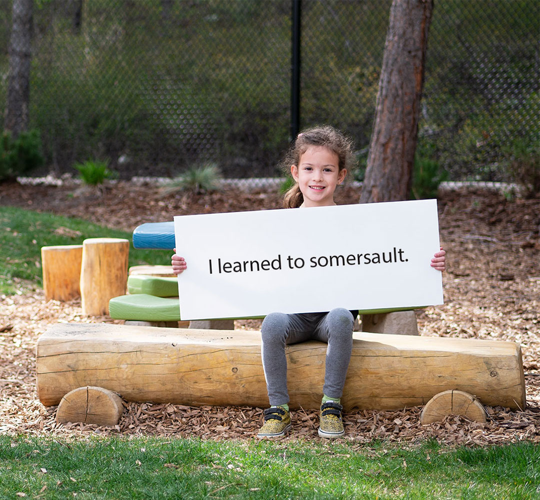 AR 2021 FA Teaser 6 year old girl sitting on a bench smiling and holding a sign that reads 'I learned to somersault.' There are trees and a yellow slide in the distance.