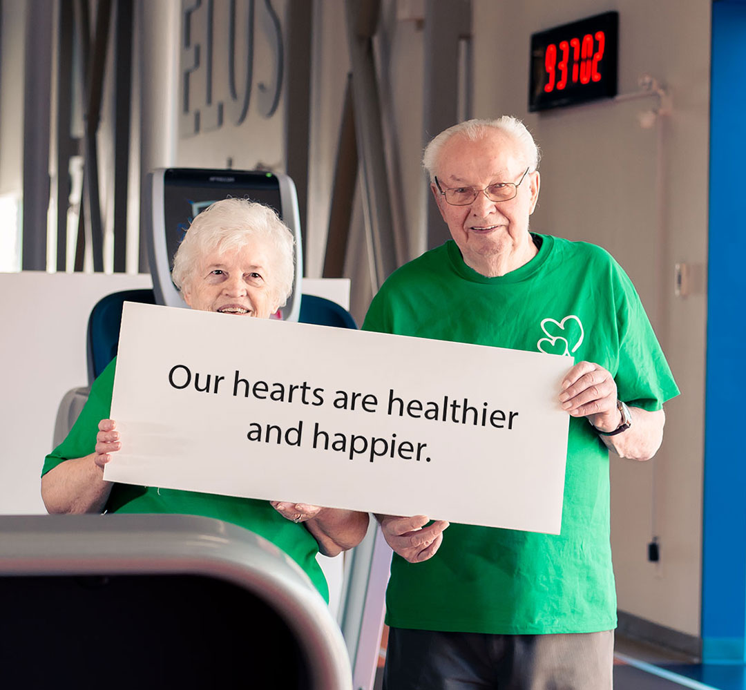 AR 2021 Specialty Health Teaser An elderly couple in green shirts smiling and holding a sign that reads 'Our hearts are healthier and happier.' There is a gym in the background.