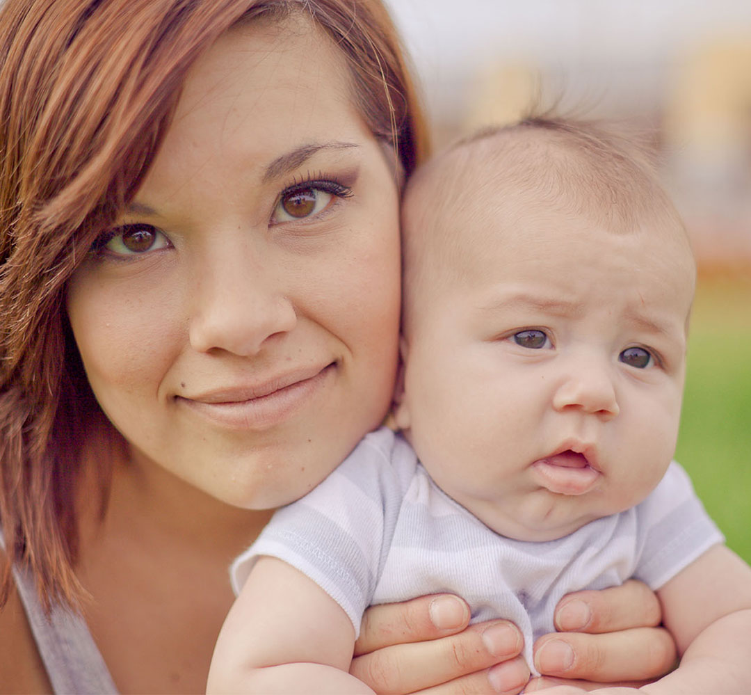 YPP Teaser A close up photo of a young mother with brown eyes and dark red hair holding a three month old baby in a white shirt.