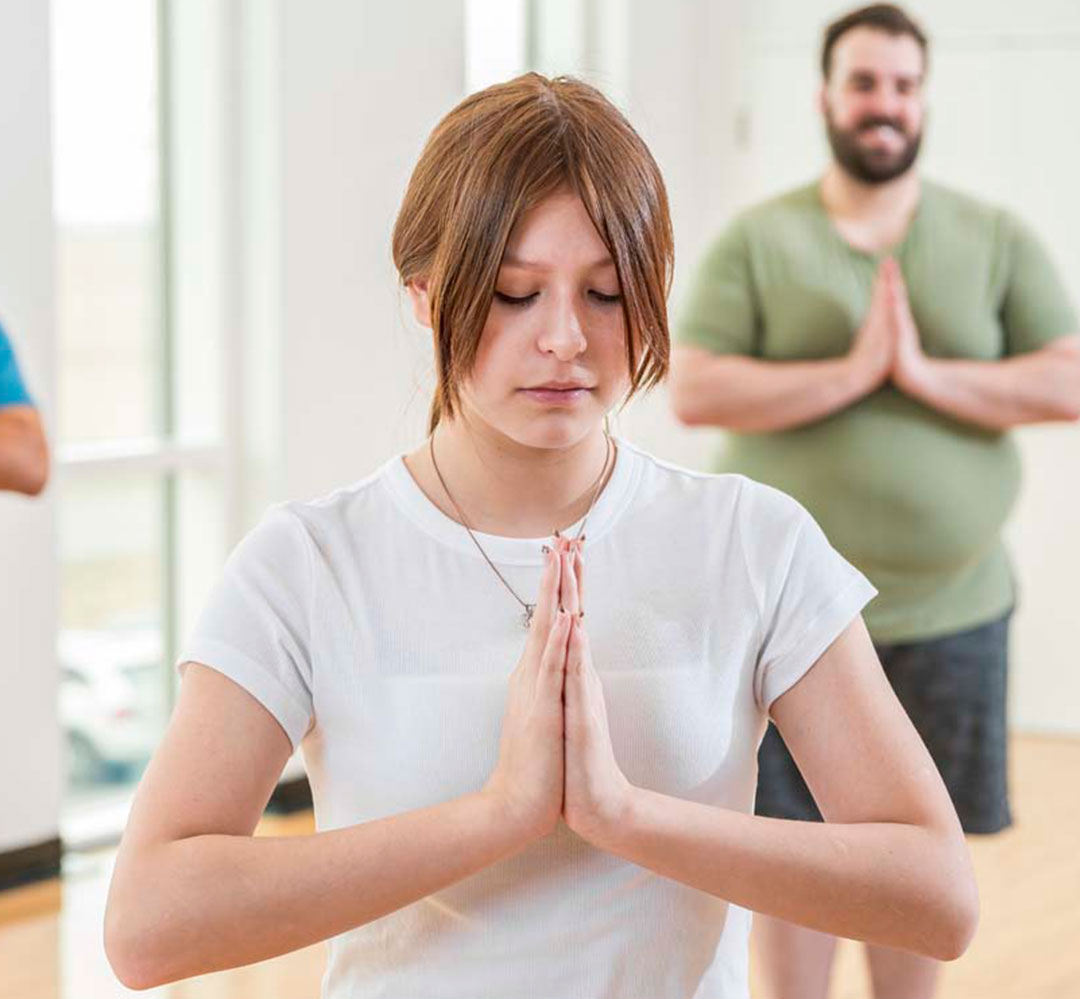 Meditation Course Teaser A young woman with auburn hair in a ponytail stand in a yoga pose with her hands together at her chest. She is in a class in a gym with people behind her.
