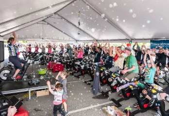  An outdoor Cycle class is under a large tent with dozens of participants dressed up brightly and cheering. There are bubbles blowing around the class and two children dancing in the foreground with one Cycle instructor on the a stage to the left leading the class. 