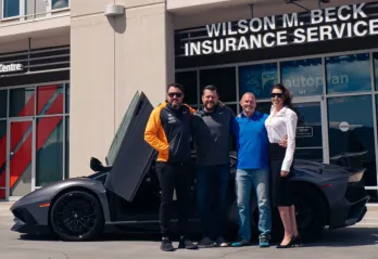 Three men and one woman are standing in front of a high-end grey sports car with suicide doors open. They have their arms around one another and are smiling at the camera. The photo is taken outside on a sunny day with offices behind them. 