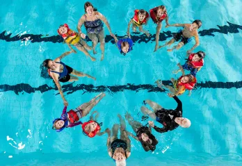 15 community members of all ages from senior to child, holding hands in a pool and looking up at the camera. Their bodies are forming the shape of a heart. 