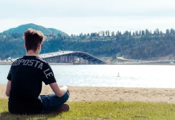 A young man with short dark hair, jeans, sun glasses and a black t shirt is sitting cross legged looking out at a beach. The photo is taken behind him and he is looking out at the water, with a bridge full of cars crossing the lake in the distance. 