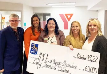 5 professionals of various ages in business clothing are standing in a YMCA office, holding a large donation cheque and smiling at the camera. 
