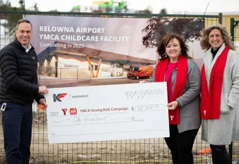 A man in a black jacket, and 2 women in grey winter coats and red YMCA scarves are all standing smiling at a camera holding a large cheque. In the background there is a construction site with a sign on the fence that reads ‘Kelowna Airport YMCA Childcare facility’.