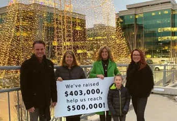 Four adults and one child are standing outside in front of an enormous Christmas tree made of Christmas lights. They are all smiling at the camera and holding a sign that reads 'We've raised $403,00, help us reach $500,000!'. 