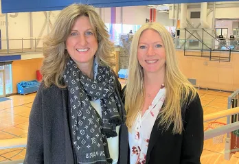 Two blonde women in professional clothing are standing in front of a gymnasium and smiling at the camera. 