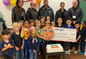 Five adults are standing in a classroom smiling at the camera. In front of them a group of 15 children aged 5-7 are smiling and holding a large donation cheque made out to the YMCA.