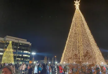 An enormous Christmas tree is lit up with all white lights  in the nighttime, as a crowd gathers around the base.  There are office buildings in the background that reflect the tree. 