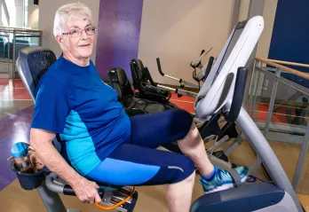 A woman with glasses, short white hair, and blue workout clothes is sitting on a laid back cycle machine in a gym and smiling at the camera. 