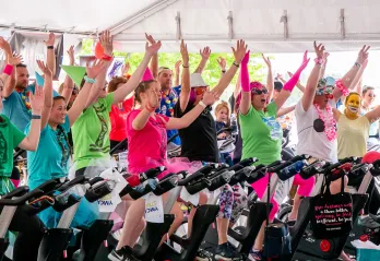 A group of 30 people are dressed up in costumes and bright colors, and sitting on stationary bikes in an outdoor class. They are all smiling and cheering and holding their hands up in the air. 