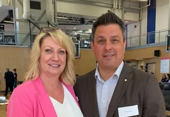 A professional woman and man are standing together smiling at the camera with an event taking place behind them in a gymnasium. 