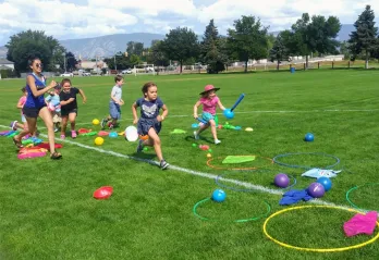 A female YMCA camp leader runs through an outdoor obstacle course with 6 children, aged 7-9, outside on a sunny day. 