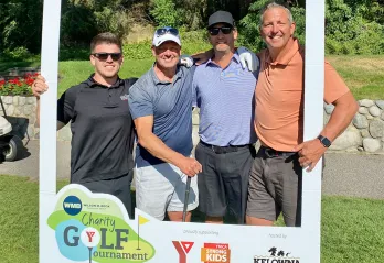 Four men in golf attire are holding clubs and smiling at the camera on a golf course.
