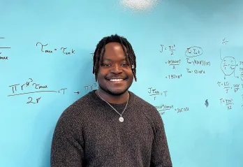 A young man with short black thin dreadlocks in a brown sweater is smiling at the camera in front of a teal board with mathematical equations scribbled on it.
