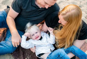 A mother and folder are holding their 2 year old boy and looking down at him as they sit on a beach. The little boy has blue eyes, blonde hair, and is smiling up at the camera.