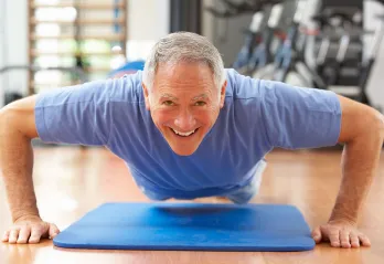 An older gentleman with grey hair and a blue t shirt is hovering in a pushup over a blue mat in a gym, and smiling at the camera. 