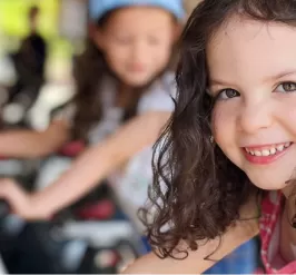 A young girl is sitting on a cycle bike and smiling at the camera as an outdoor cycle class takes place behind her