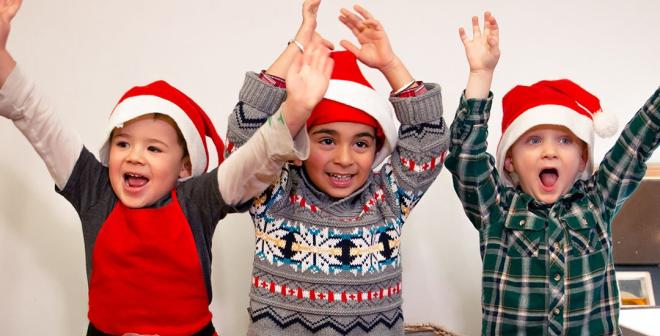 Three children wearing Santa hats raise their arms happily