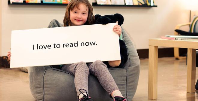 Young girl sitting back in a bean bag chair smiling and holding a large white sign that reads 'I love to read now.' There is a classroom with books in the background. 