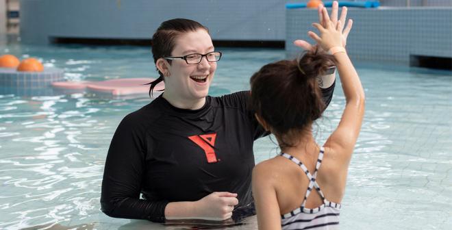 A young female swim lesson instructor with glasses and a black rash guard with a red YMCA logo is smiling at and high fiving a 9 year old girl. They are both kneeling in a pool facing one another.  
