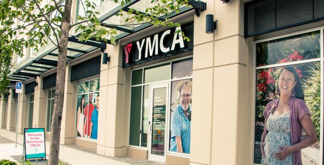 The YMCA front doors on the bottom floor of a large building. There are trees outside on the sidewalk and large photos of people smiling inlaid into the windows so you can't see inside.    