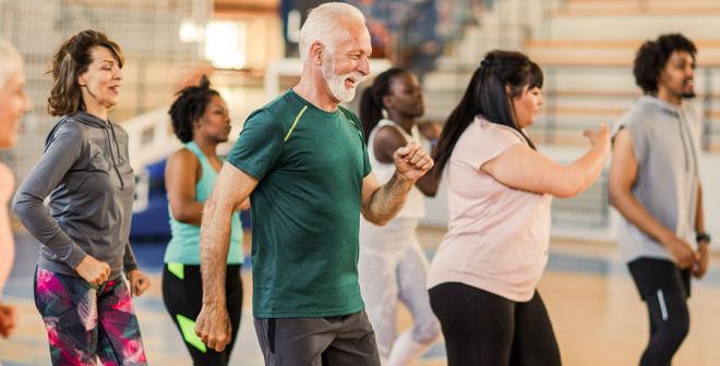 Seven adults in a group fitness class all mid step with arms swinging. There is an older gentleman in front with a green shirt and grey hair smiling. 