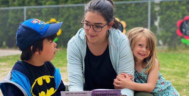 A young adult with glasses and dark hair in a ponytail reading a book to 2 4-year old children outside in a park. One boy with dark hair and a hat is talking to her and one girl with blonde hair is hugging her arm and smiling at the camera.