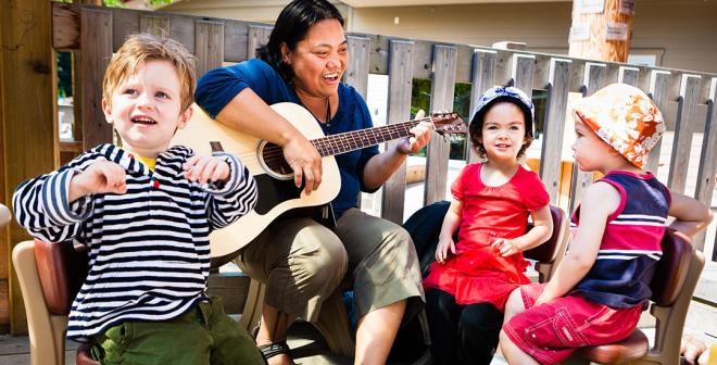 A woman with dark hair and a guitar in hand is sitting outside on a deck in the sun. She is smiling at the three 4-year old children that are sitting around her. 