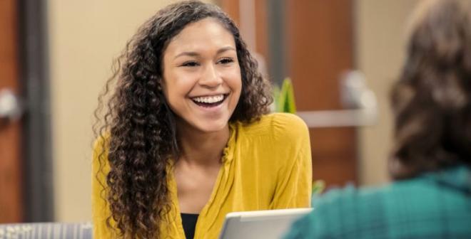 A young woman with long brown curly hair in a yellow blouse is holding a tablet and smiling at someone off camera. 