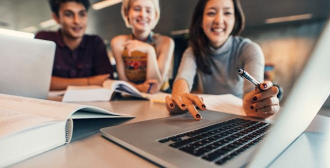 Three professionally dressed young adults are sitting at a table covered in books and smiling at a laptop. One of them is controlling the mouse. 