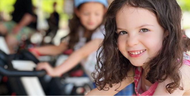 A young girl is sitting on a cycle bike and smiling at the camera as an outdoor cycle class takes place behind her