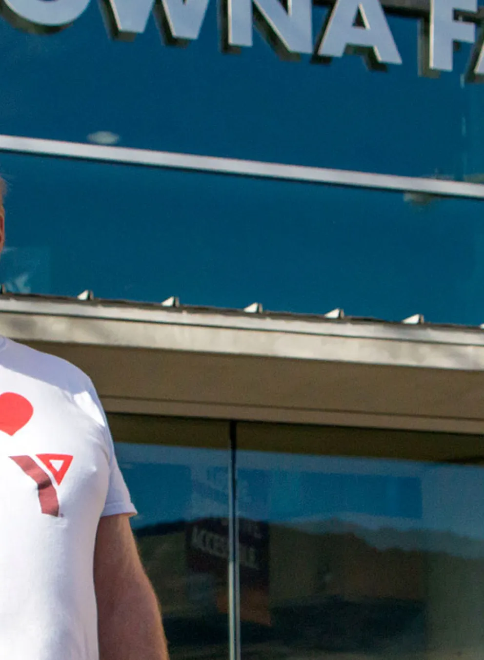 A man with glasses and a grey goatee is standing in front of the Kelowna Family Y and smiling at the camera. He’s wearing a white shirt that says ‘I love my Y’.