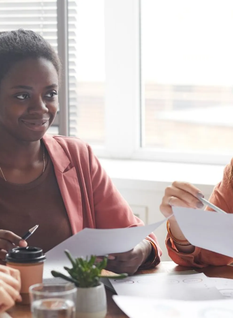 Four young women in professional clothing are sitting around a workplace table, holding pens and papers as they smile at one woman who is speaking.