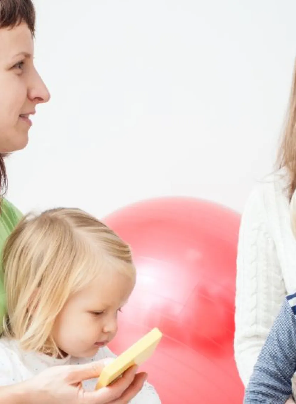 A group of young moms are sitting in a play room with their 3 year old children on their laps. They are smiling and listening to a speaker who is off screen.