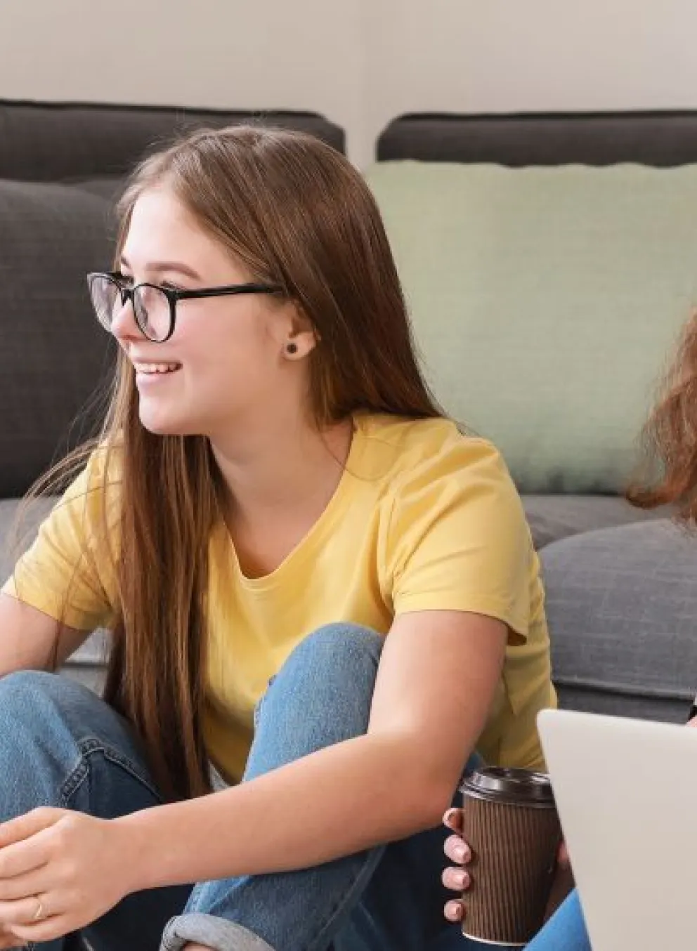 Four teens are sitting in a group centre. They are smiling at each other. Two females are holding a phone and a coffee, and two males are holding a laptop and books.