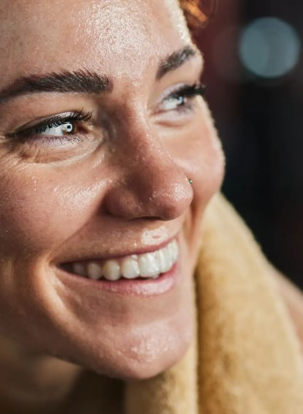 A middle aged woman with dark curly hair in a pony tail and seat towel around her neck is sitting in a gym. She's covered in sweat and smiling off to the side.