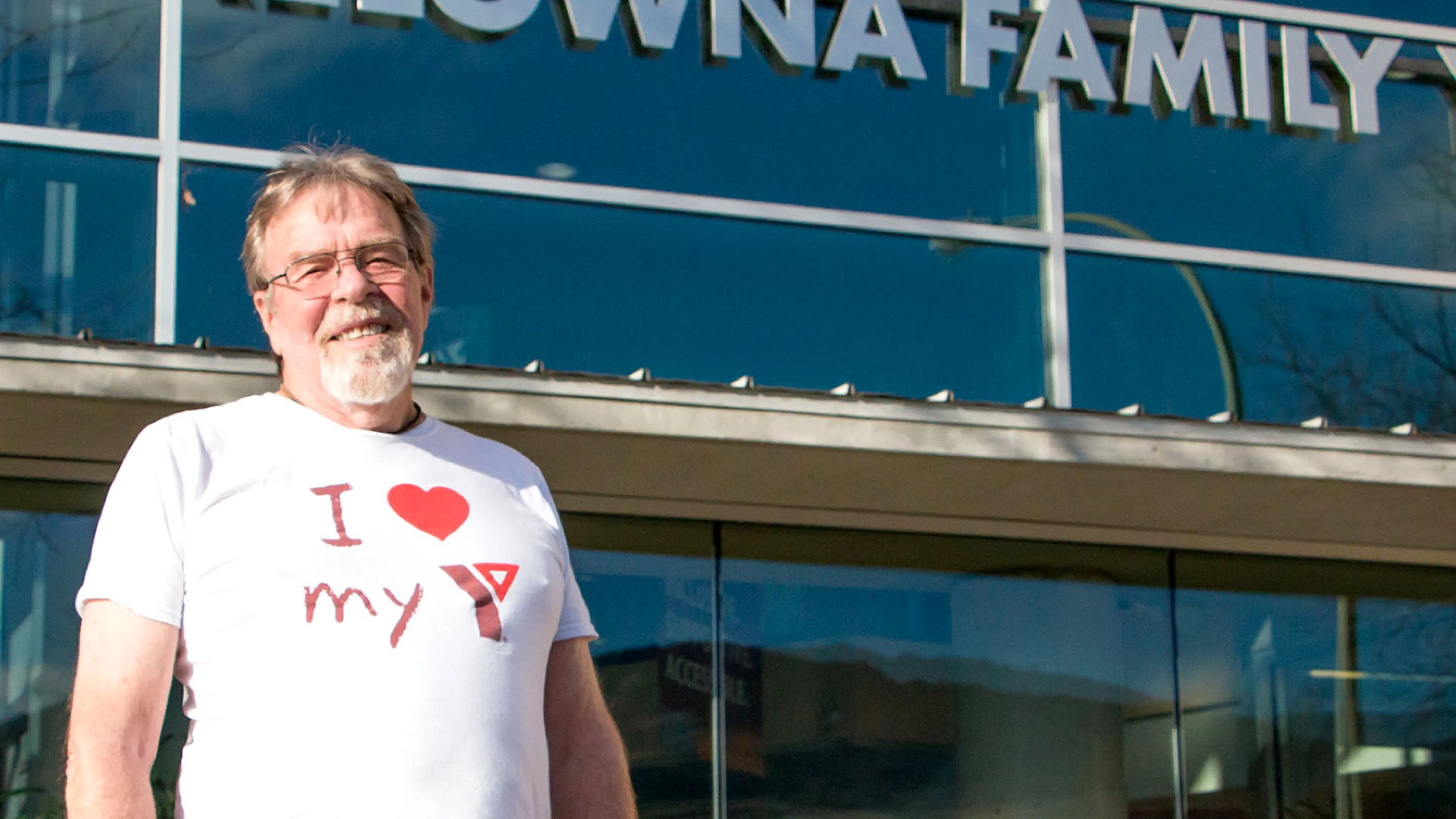 A man with glasses and a grey goatee is standing in front of the Kelowna Family Y and smiling at the camera. He’s wearing a white shirt that says ‘I love my Y’.