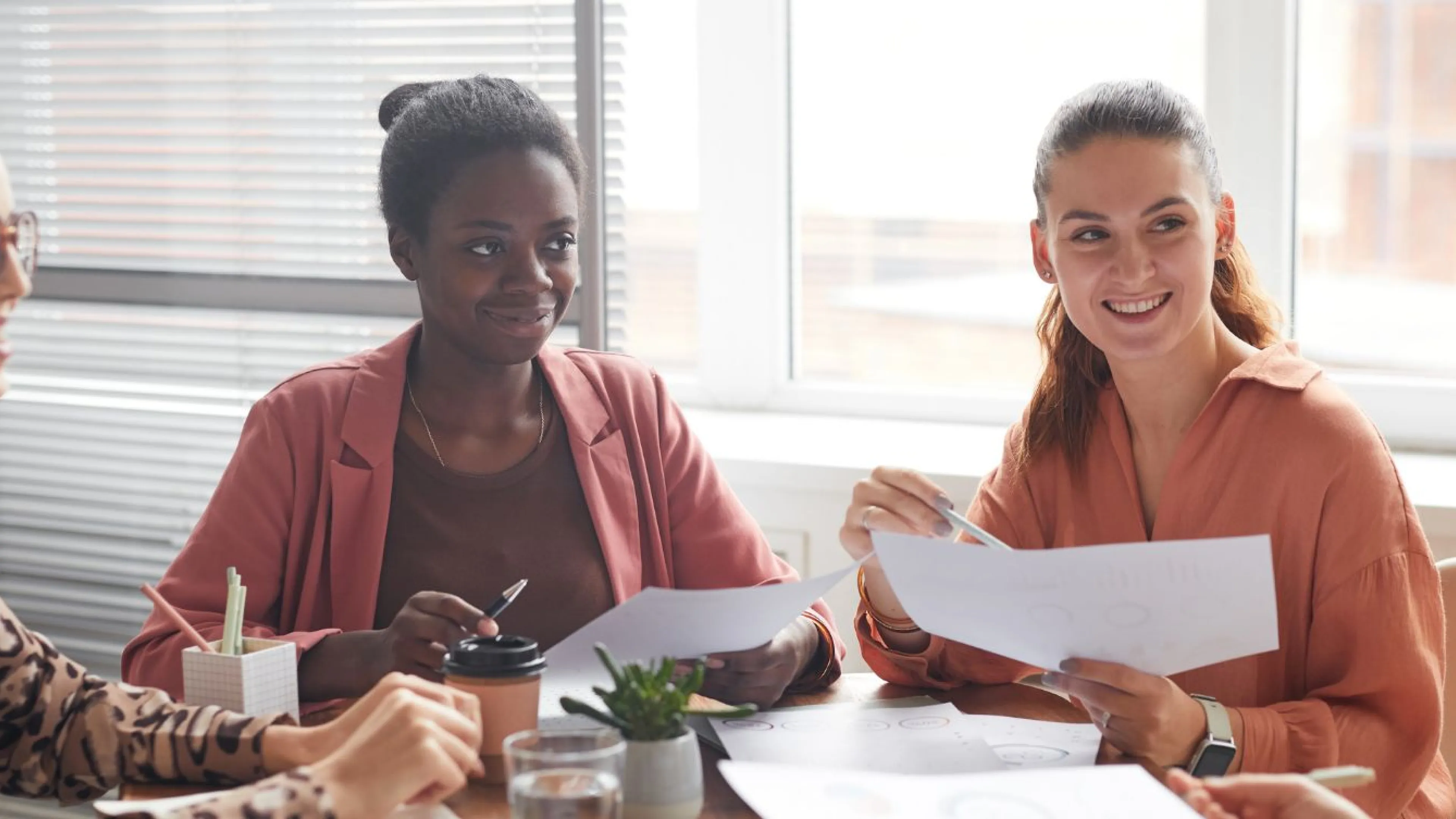 Four young women in professional clothing are sitting around a workplace table, holding pens and papers as they smile at one woman who is speaking.