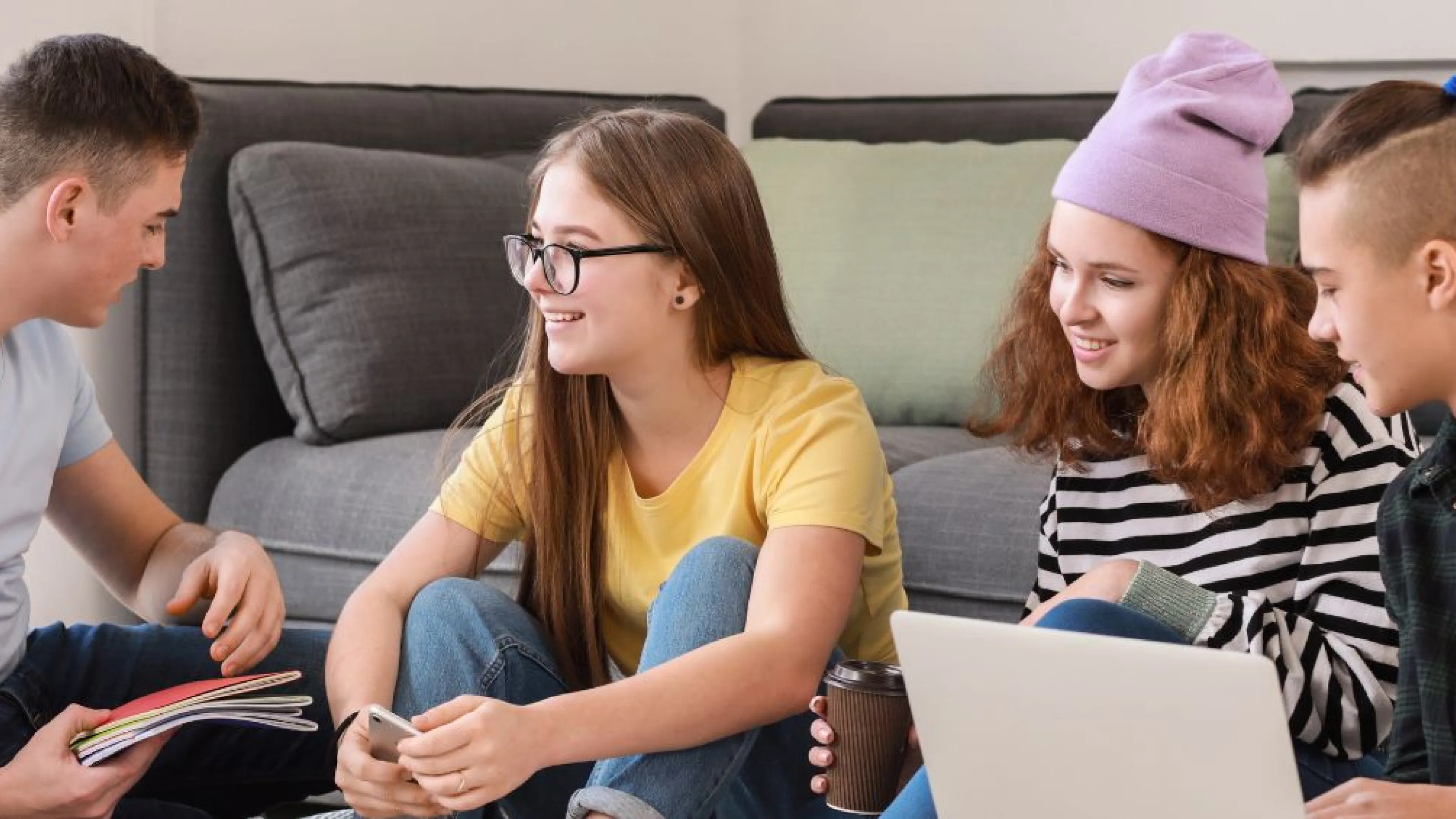 Four teens are sitting in a group centre. They are smiling at each other. Two females are holding a phone and a coffee, and two males are holding a laptop and books.