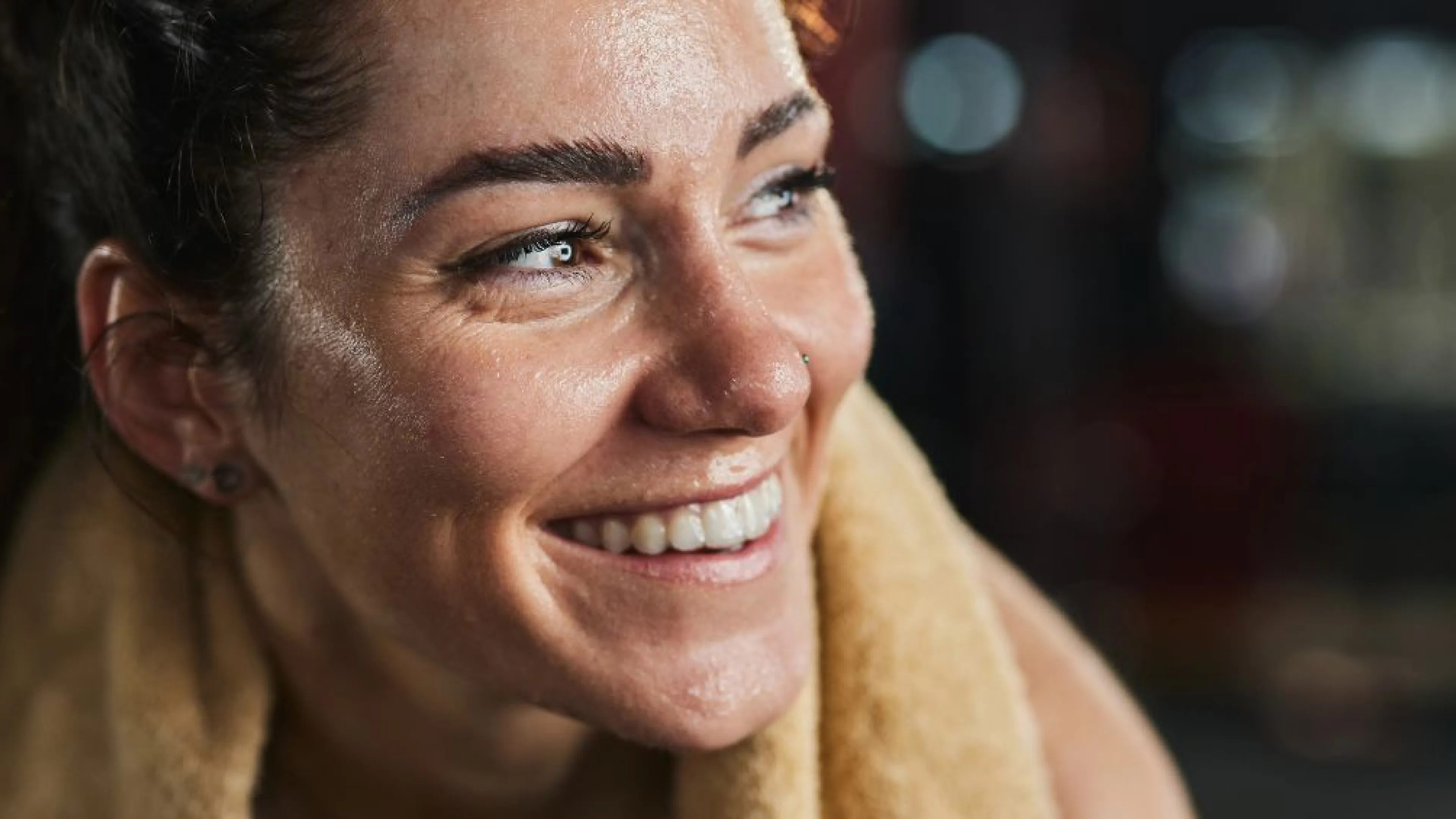 A middle aged woman with dark curly hair in a pony tail and seat towel around her neck is sitting in a gym. She's covered in sweat and smiling off to the side.