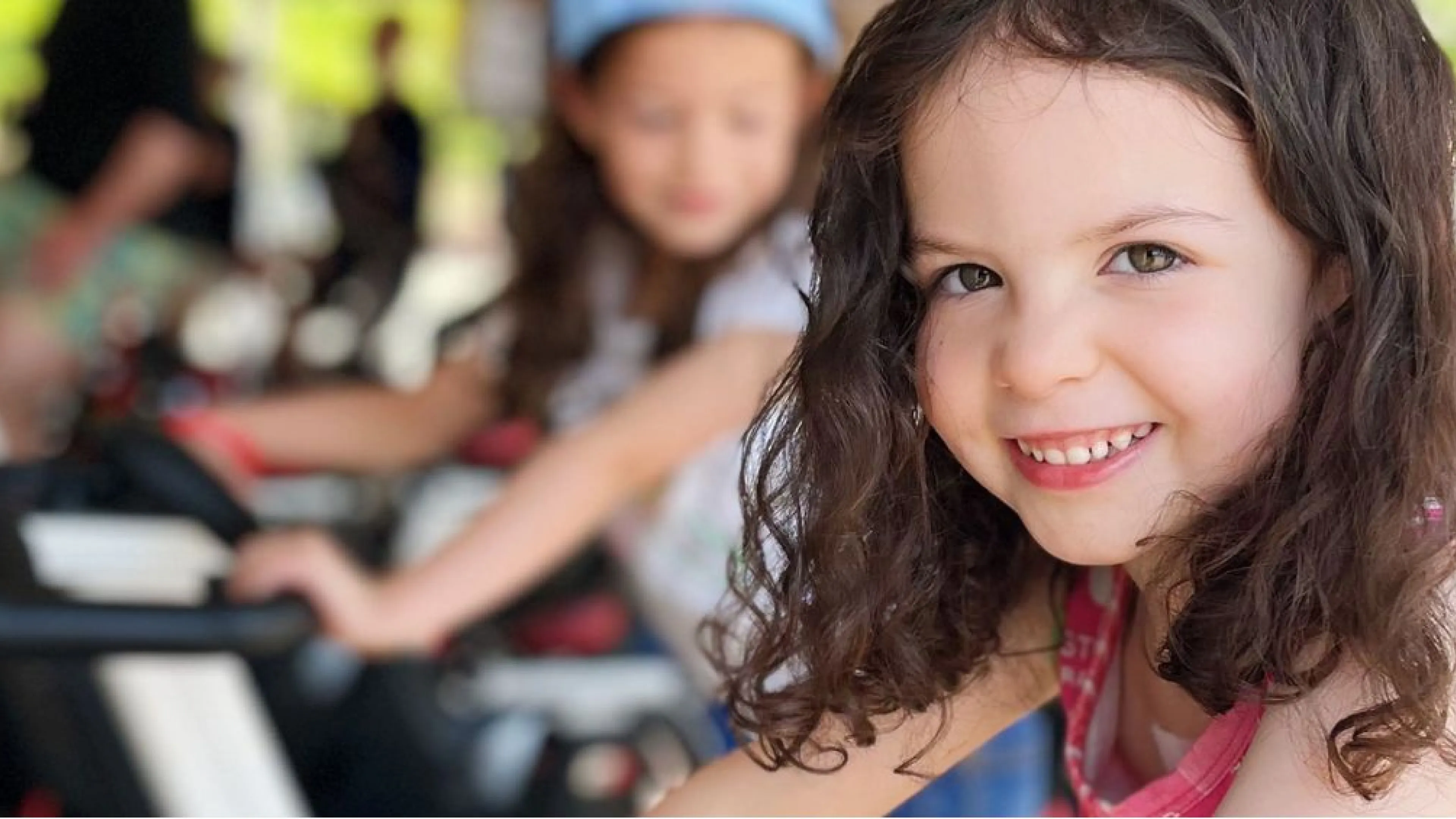 A young girl is sitting on a cycle bike and smiling at the camera as an outdoor cycle class takes place behind her