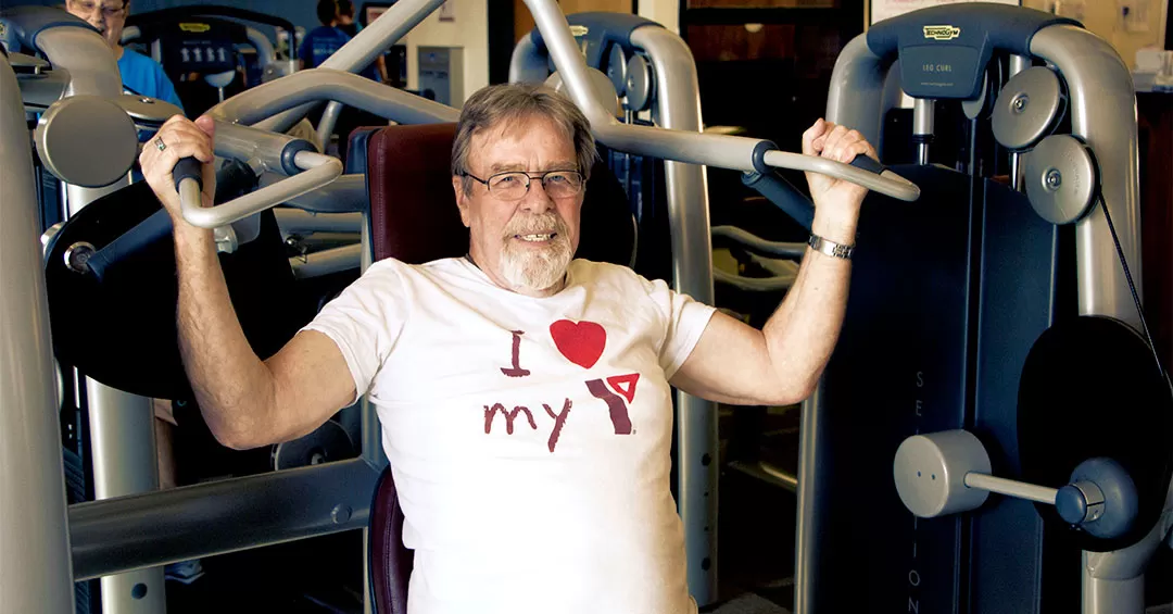 A man with glasses and a grey goatee is sitting at a weights machine in a gym with his arms on the handles and smiling at the camera. He’s wearing a white shirt that says ‘I love my Y’.