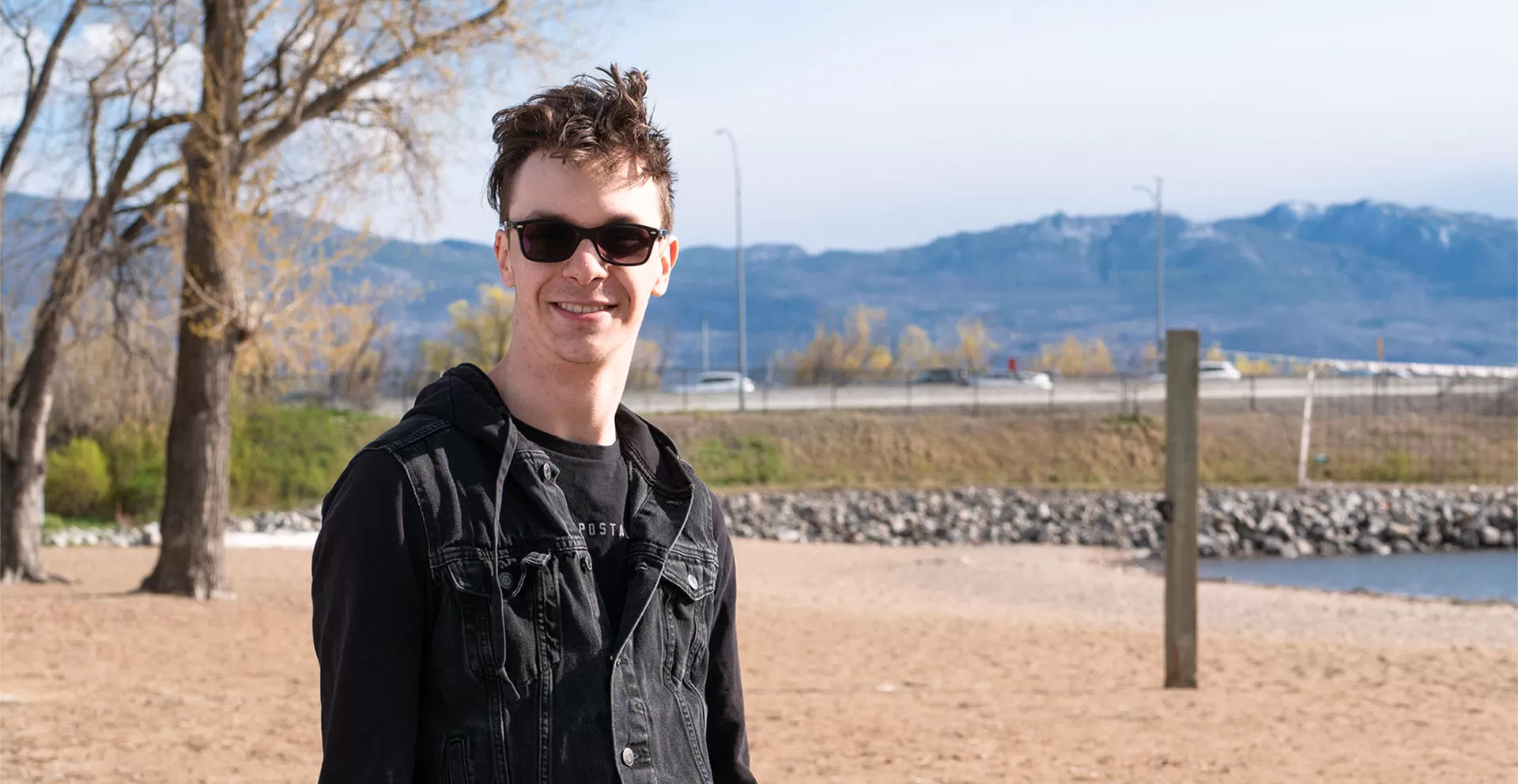 A young man with short dark hair, sunglasses, a black t shirt and black jean jacket is outside on a beach smiling at the camera on a sunny day.  