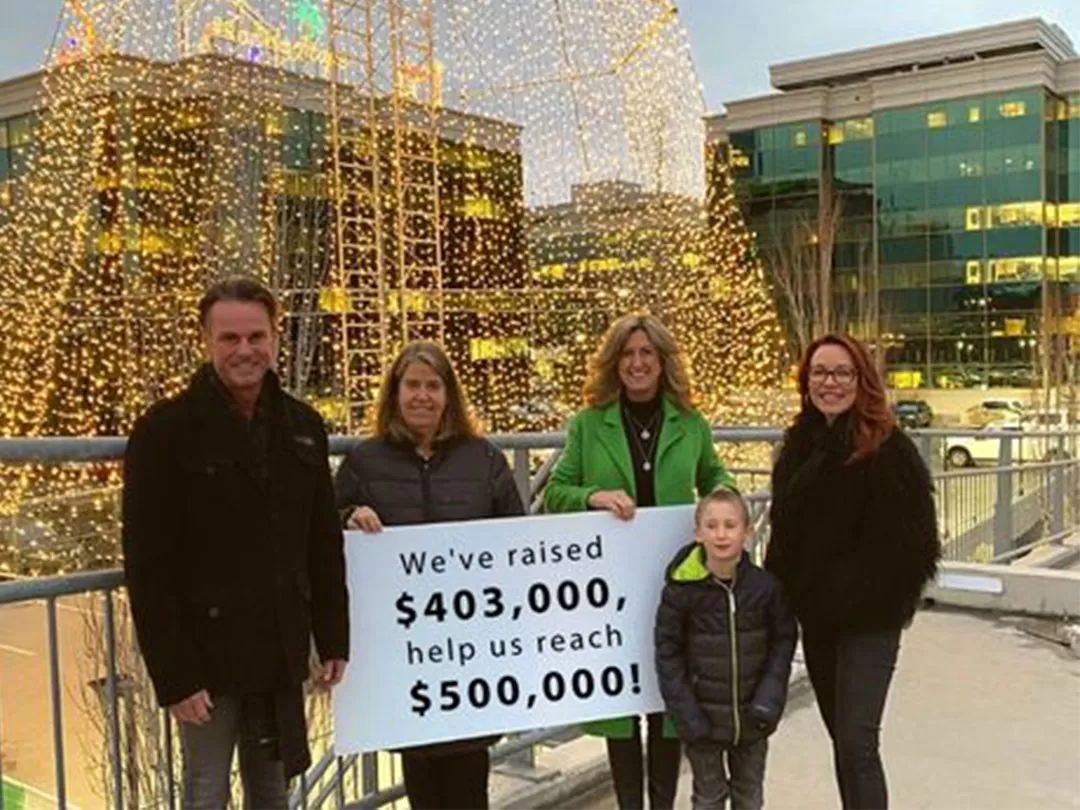 Four adults and one child are standing outside in front of an enormous Christmas tree made of Christmas lights. They are all smiling at the camera and holding a sign that reads 'We've raised $403,00, help us reach $500,000!'. 