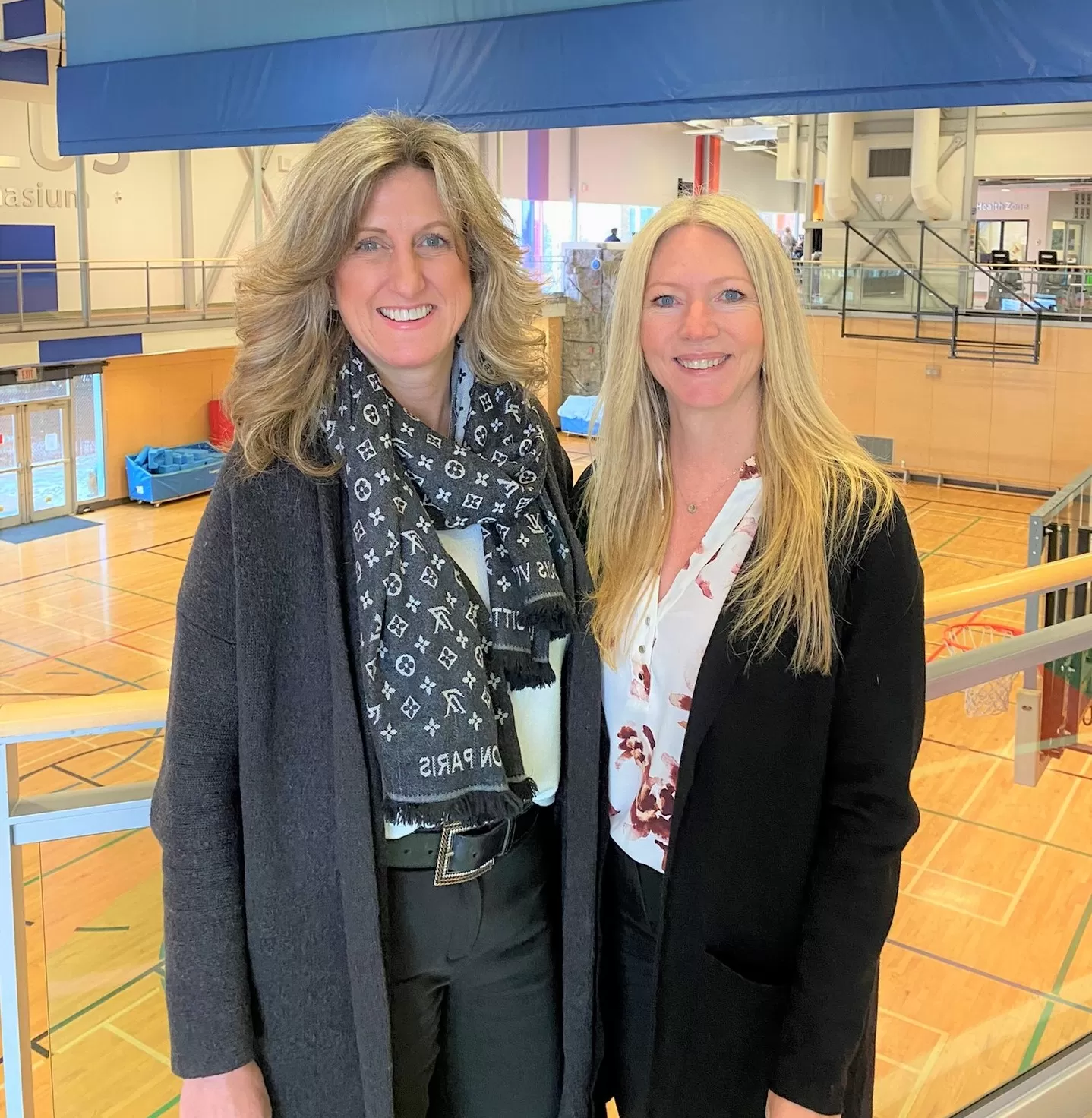 Two women in professional clothing and blonde hair are standing in front of a gymnasium and smiling at the camera. 