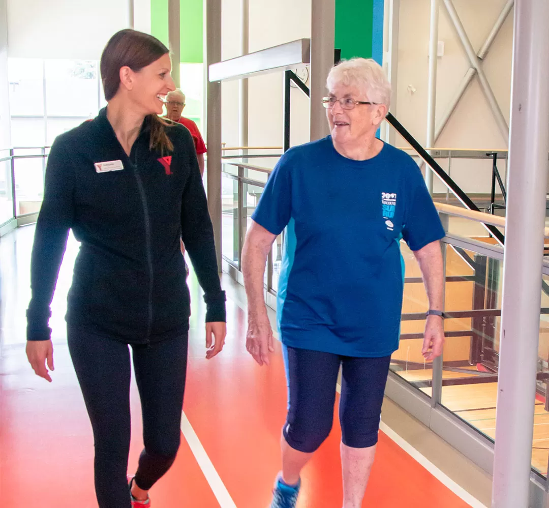 Two women are wearing workout clothes and walking an indoor track at a gym. They are smiling at each other, one is a YMCA staff member with dark hair, the other is a member with white hair. 